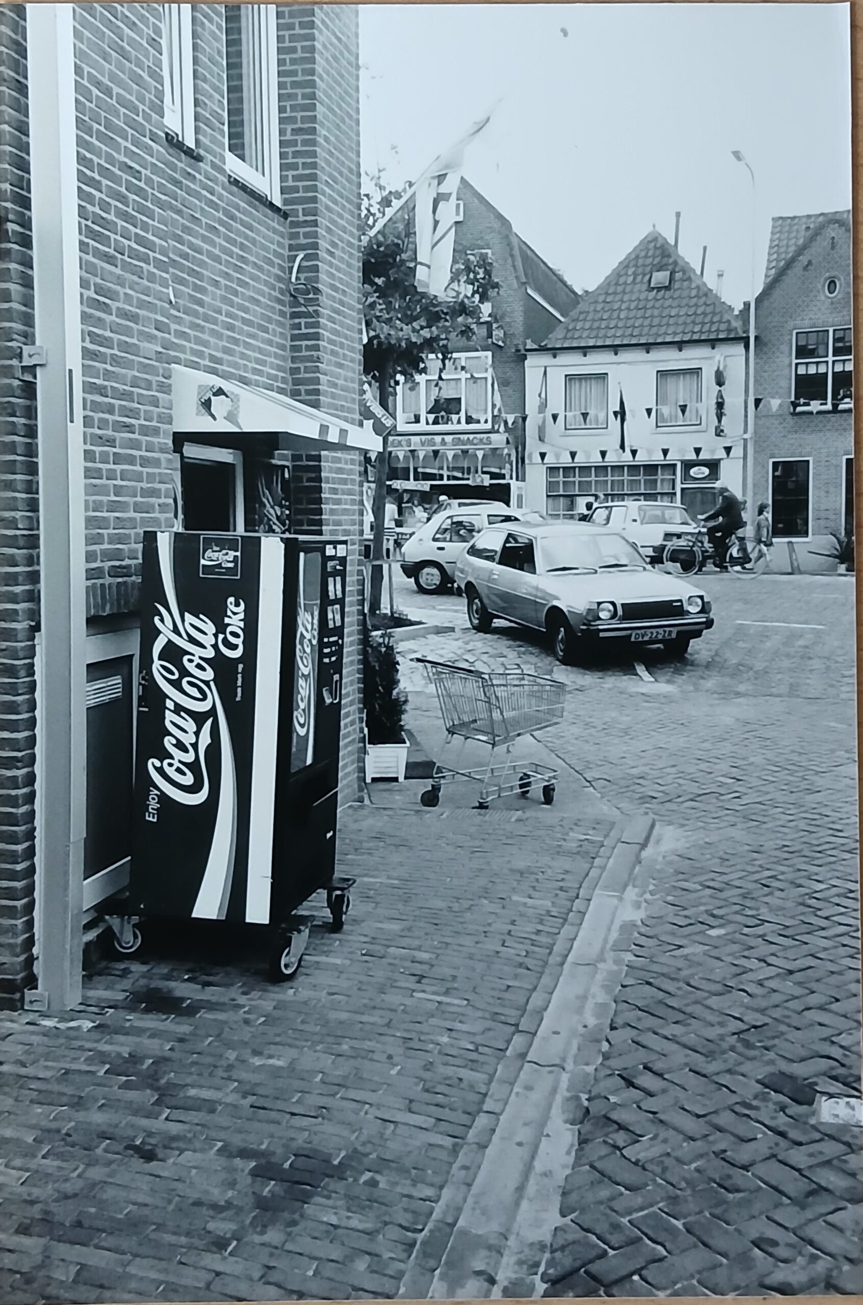 alte Schwarzweiß-Postkarte einer europäischen Straße aus den 60er/70ern, mit Auto im Vordergrund und typischem Straßenleben im Hintergrund.