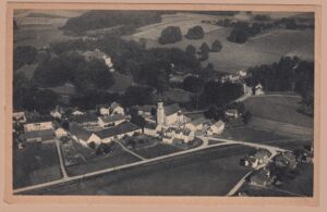 Schwarz-weiße Flugzeugaufnahme des oberbayerischen Dorfes Egglkofen (Obb.) aus der Vogelperspektive, um 1920: Zu sehen sind die Kirche mit schlankem Zwiebelturm, der angrenzende Friedhof, traditionelle Bauernhöfe und eine Bahnlinie inmitten sanfter Hügellandschaft. Herausgegeben als „FLUG-POSTKARTE" der Flugphoto Verlags-Ges. München unter Nr. 280 – ein frühes Luftbild und postgeschichtlicher Beleg der Nachkriegszeit, gelaufen 1920 nach Mülhausen im Elsass.