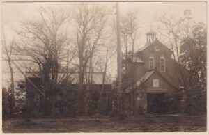 Sepiagetönte Real-Photo-Postkarte (RPPC) einer protestantischen Missionskirche in ländlichem Japan, Meiji- bis frühe Showa-Zeit (ca. 1890er–1930er Jahre): Der holzverkleidete Bau verbindet neugotisch-westliche Elemente – schmale gotische Spitzbogenfenster, giebelbekrönter Eingangsvorbau und ein kleines offenes Glockentürmchen (Dachreiter) mit spitzem Helm auf dem Dachfirst – mit typisch japanischen Konstruktionsmerkmalen wie leicht ausgestellten Dachkanten und Holz-Putzbauweise. Links steht ein verbundenes Nebengebäude, vermutlich Pfarrhaus, Schulraum oder Gemeinschaftssaal einer christlichen Missionsstation; kahle Winterbäume rahmen die Szene, ein hölzerner Telegrafenmast ragt zentral empor. Die Rückseite bestätigt die japanische Herkunft eindeutig: Die Karte wurde auf „Oriental OK Photo Papers" der Oriental Photo Industrial Co., Ltd. (Tokio, gegründet 1919) abgezogen – ein Fotopapier, das hauptsächlich in den 1920er bis 1930er Jahren (Taishō- bis frühe Shōwa-Zeit) für echte Fotopostkarten verwendet wurde. Ungelaufen und unbeschrieben, dokumentiert diese RPPC in warmer Sepiatönung ein seltenes Zeitzeugnis früher Missionsarchitektur in Japan – ein Sammlerstück für Freunde von RPPC-, Japan- und Architektur-Motiven.