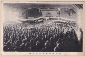 Schwarz-weiße japanische Foto-Ansichtskarte aus der Vorkriegszeit (Taishō bis frühe Shōwa) der Tōeisha-Ehrungszeremonie (東英社勤続表彰式式場) für langjährige Betriebszugehörigkeit. Hunderte Angestellte sitzen in einer rustikalen Holzhalle vor einer festlich geschmückten Bühne mit dunklem Zeremonienvorhang (maku), Würdenträgern und hängendem Kanji-Banner. Ein eindrucksvolles Zeugnis japanischer Unternehmenskultur, das Loyalität, Mitarbeiterbindung und sichtbare Wertschätzung dokumentiert – ein reizvolles Sammlerstück für Freunde von Japan-, Foto- und Firmengeschichte.