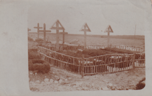 Historische Fotokarte: hölzerne Grabkreuze, drei davon mit dreieckiger Überdachung, hinter einem Zaun aus Ästen auf einer kleinen Gräberanlage in flacher Landschaft