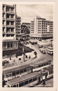 Schwarz-Weiß-Fotokarte des Berliner Alexanderplatzes, von oben aufgenommen: Straßenbahnen im Vordergrund, dahinter ein Doppeldeckerbus, Lastwagen, Autos und Passanten vor großen Geschäftshäusern und der gewölbten Bahnhofshalle.